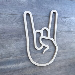 May include: A wooden cutout of a hand making the rock and roll hand gesture. The cutout is white and is mounted on a gray wood background.