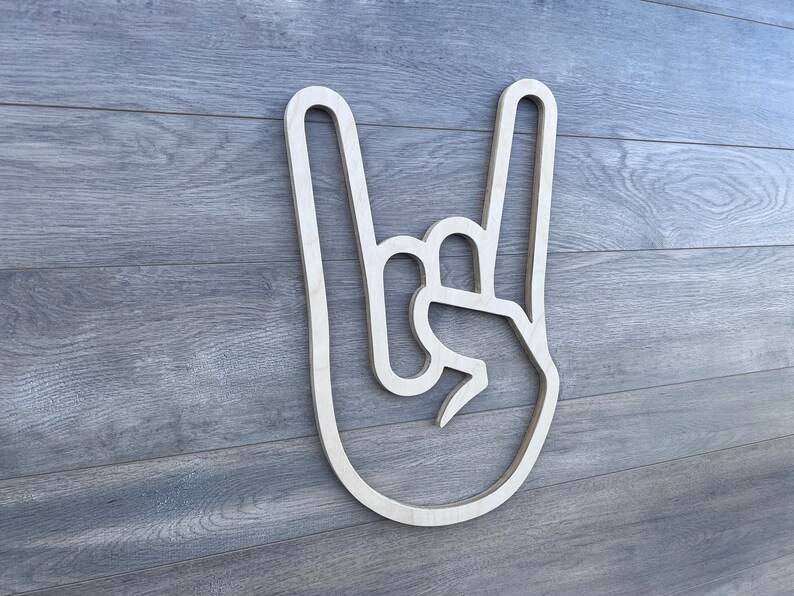 May include: A wooden cutout of a hand making the rock and roll hand gesture. The cutout is white and is mounted on a gray wood background.