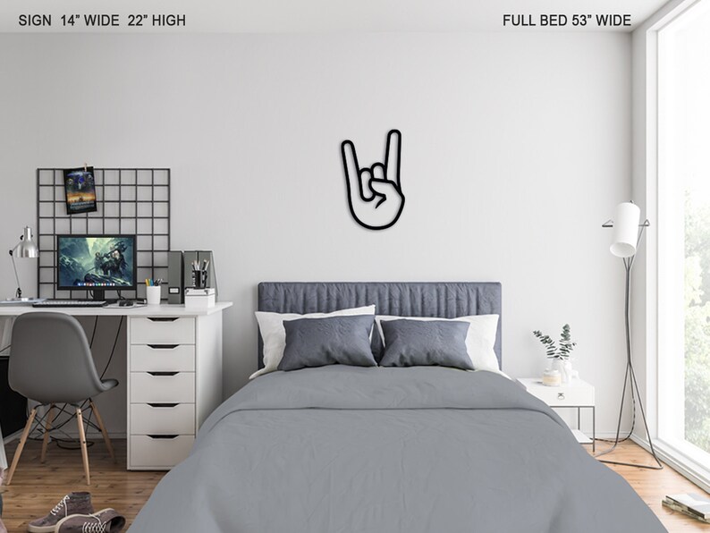 May include: Black metal wall decor in the shape of a hand making the rock and roll hand gesture. The decor is mounted on a white wall above a gray bed with white pillows. The bed is in a bedroom with a desk and chair, a lamp, and a plant.