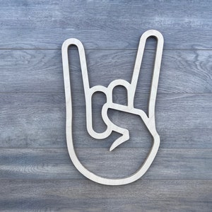 May include: A white wooden cutout of a hand making the rock and roll hand gesture. The hand is outlined in white against a wood grain background.