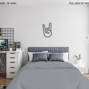 May include: A gray metal wall decor in the shape of a hand making the rock and roll hand gesture. The decor is mounted on a white wall above a gray upholstered bed with white pillows. The bed is in a bedroom with a desk and chair in the background.