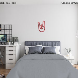May include: Red metal wall decor in the shape of a hand making the rock and roll hand gesture. The decor is mounted on a white wall above a gray bed with white pillows. The bed is in a bedroom with a desk and chair, a lamp, and a plant.