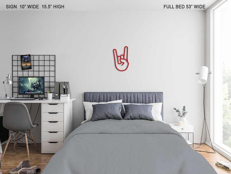 May include: Red metal wall decor in the shape of a hand making the rock and roll hand gesture. The decor is mounted on a white wall above a gray bed with white pillows. The bed is in a bedroom with a desk and chair, a lamp, and a plant.