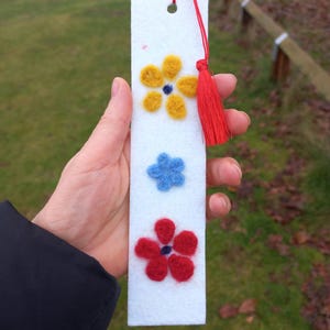 Op de afbeelding: Witte vilt boekenlegger met een rode kwast. De boekenlegger heeft drie vilt bloemen in geel, blauw en rood.