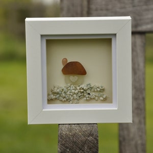 May include: A white framed picture with a small house made of sea glass and pebbles. The house has a heart-shaped window and is surrounded by small white pebbles.