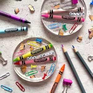 May include: Two clear resin coasters with colourful crayon pieces, paper clips and push pins embedded inside. The coasters have the names "Ellen" and "Mrs. Smith" spelled out with white letter beads on top of the crayons.