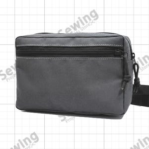 Puede incluir: Un estuche utilitario rectangular de color gris carbón que presenta un bolsillo frontal horizontal con cremallera, herrajes negros y costuras blancas en contraste. El bolso está fabricado con tela tejida resistente e incluye una correa negra.