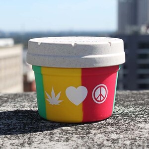 Puede incluir: Un pequeño contenedor redondo blanco con un diseño verde, amarillo y rojo. El contenedor tiene un símbolo de paz blanco, un corazón blanco y una hoja de marihuana blanca impresa.