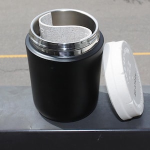 May include: A black insulated container with a stainless steel interior and a white lid. The lid is off to the side. Inside the container is a light gray, curved insert. The container is sitting on a gray surface.