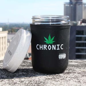 Könnte beinhalten: Schwarzes Glas mit einem durchsichtigen Glasdeckel und einem weißen Deckel. Auf dem Glas steht das Wort "CHRONIC" in Weiß mit einer grünen Cannabisblatt-Grafik. Auf dem Deckel steht das Wort "RESTORE". Ein Warnhinweis für Eltern ist ebenfalls auf dem Glas aufgedruckt.