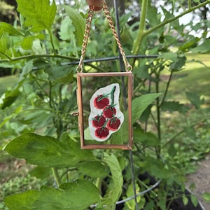 May include: A rectangular copper-toned metal frame with a glass front and back, containing a small piece of white fabric embroidered with a cluster of five red tomatoes on a green vine, suspended by a matching copper-toned metal chain.