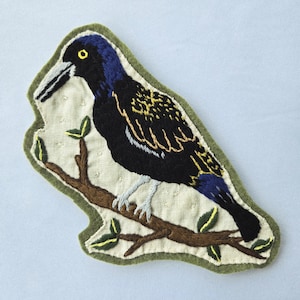 May include: An embroidered fabric patch featuring a black bird with iridescent blue and gold wing details perched on a brown branch. The bird has a yellow eye and a light-colored beak. The patch has a light beige background and a green border.