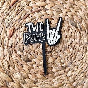 May include: Black and white cake topper with the words "TWO PUNK" and a skeleton hand making a rock and roll gesture. The topper is on a black stick, perfect for a birthday cake.