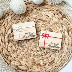 May include: Two small, rectangular wooden gift tags with the words "merry christmas" in a script font. One tag has a white bow and string, the other has a red bow and string. The tags are on a woven, circular placemat.