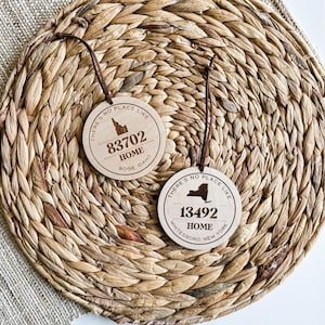 May include: Two wooden ornaments with the text "There's no place like home" and the state outlines of Idaho and New York. The Idaho ornament has the text "83702 Boise, Idaho" and the New York ornament has the text "13492 Whitesboro, New York".