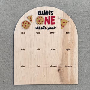 May include: A wooden milestone board with the text "ELIJAH'S ONE whole year". The board has pizza slice and whole pizza graphics. It includes numbered boxes from one to twelve for monthly milestones.