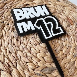 May include: Black and white cake topper with the text "BRUH I'M 12" in a bold, blocky font.