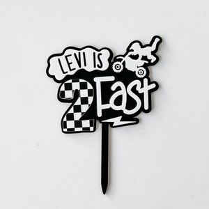 Layered acrylic 'Two Fast' second birthday cake topper with checkered number and dirt bike detail in black and white, on a wicker surface