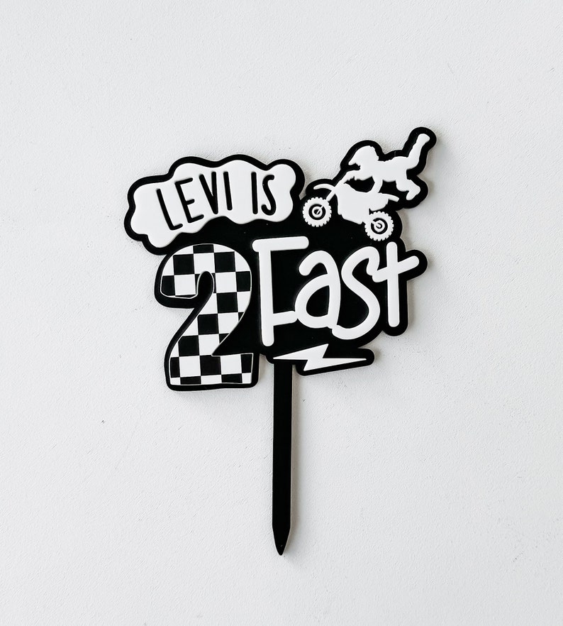 Layered acrylic 'Two Fast' second birthday cake topper with checkered number and dirt bike detail in black and white, on a wicker surface