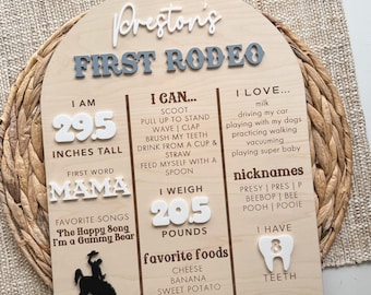 Könnte beinhalten: Ein bogenförmiges Meilenstein-Schild aus Holz mit der Aufschrift Preston's FIRST RODEO und 3D-Schriftzug. Es listet Daten wie 29.5 INCHES TALL, 20.5 POUNDS, das erste Wort MAMA sowie Lieblingslieder, Speisen, Spitznamen und Aktivitäten auf.
