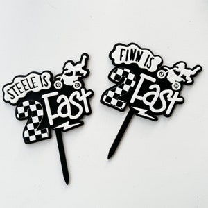 Pair of layered acrylic 'Two Fast' second birthday cake toppers with checkered flag and dirt bike motif in black and white, on black acrylic stakes