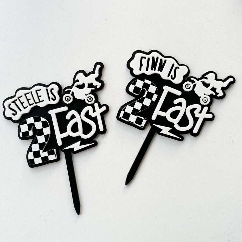Pair of layered acrylic 'Two Fast' second birthday cake toppers with checkered flag and dirt bike motif in black and white, on black acrylic stakes