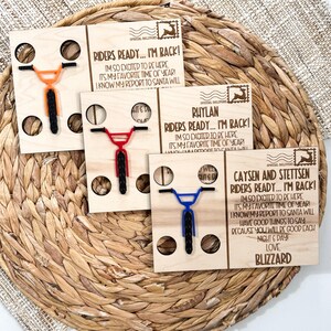 May include: Wooden Christmas cards with bicycle designs in orange, red, and blue. Each card has a bicycle cutout and text that reads "Riders Ready... I'm Back!" and personalized names. The cards are on a woven basket.
