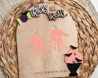 May include: Wooden Halloween sign with the text "No Tricks All Treats" and "Halloween 2025". The sign features two handprint pumpkins and a cupcake with bats. The sign is on a woven placemat.