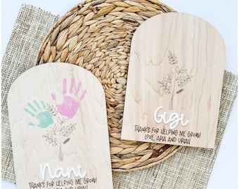 Può includere: Due targhe in legno con la parte superiore ad arco. Una presenta impronte di mani in menta e rosa, l'altra il nome "Gigi". Entrambe hanno disegni floreali e il testo "THANKS FOR HELPING ME GROW! LOVE, ARIA AND UPIAH."