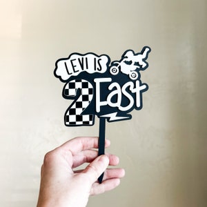 Black and white layered acrylic 'Two Fast' cake topper with dirt bike motif, displayed on a light neutral background