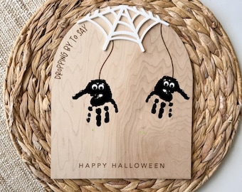 May include: A wooden Halloween decoration featuring two black handprint spiders hanging from a white spiderweb. The sign reads "HAPPY HALLOWEEN" and "DROPPING BY TO SAY". The background is a woven, circular placemat.