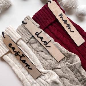 May include: Three white knitted stockings with wooden tags attached. The tags are labeled "Mom", "Dad", and "Easton".