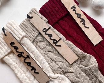 May include: Three white knitted stockings with wooden tags attached. The tags are labeled "Mom", "Dad", and "Easton".