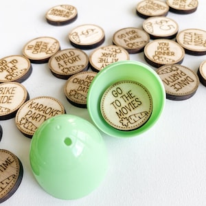 May include: A green plastic Easter egg with a wooden circle inside that says "Go to the Movies". The wooden circle has a small carrot drawing on it. There are other wooden circles with different activities written on them, such as "Go out to eat", "Bike Ride", "Karaoke Party", "Build a Fort", "Hot Chocolate Date", "Park at the Bay", "Go for a Hike", "You Pick Dinner", "Dance Party", "Pizza Night", "Smores Night", "No Chores Today", "Breakfast for Dinner", and "1$".