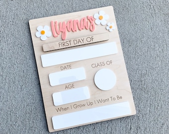 May include: A wooden sign with the name "Lilyana's" in pink lettering and white flowers. The sign has spaces to fill in the date, class, age, and what the child wants to be when they grow up.