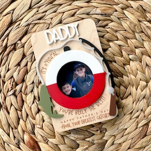 May include: A wooden Father's Day card with a photo frame shaped like a fishing bobber. The card says "Daddy, It's "Fishi"al, You're Reely the Best! Happy Father's Day -From, Your Greatest Catches".