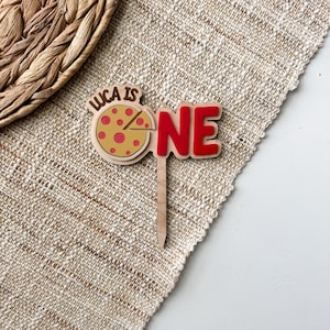 May include: A wooden cake topper with the text "LUCA IS ONE". The topper features a pizza slice design with red pepperoni and a red "ONE" text. The topper is designed for a first birthday celebration.