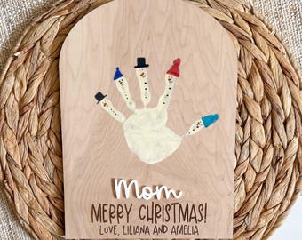 May include: A wooden plaque with a handprint painted to resemble snowmen, each finger wearing a different colored hat. The words "Mom Merry Christmas! Love, Liliana and Amelia" are written on the plaque.