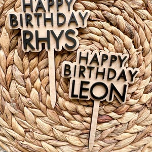 May include: Two wooden cake toppers with the words "HAPPY BIRTHDAY RHYS" and "HAPPY BIRTHDAY LEON" in black lettering. The toppers are on a light brown background.