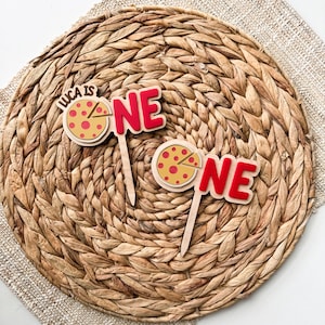 May include: Two wooden cake toppers with a pizza slice design and the word "ONE" in red. One topper also includes the text "LUCA IS". The toppers are on a woven placemat.