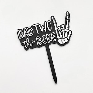 May include: Black cake topper with white text that reads "Bad Two the Bone" and a white skeleton hand making a rock and roll sign.