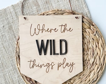 May include: Wooden sign with the phrase "Where the WILD things play" in a mix of script and bold black letters. The sign is light-colored with a brown leather hanging strap, resting on a woven mat.
