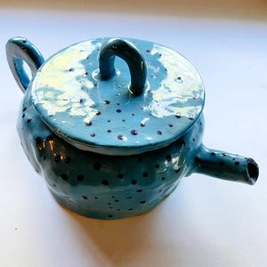 Può includere: Teiera in ceramica turchese fatta a mano con coperchio abbinato. La teiera presenta un corpo arrotondato, un manico curvo e un beccuccio. La superficie è macchiata di punti blu scuro. Il coperchio ha un manico curvo.