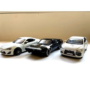 Op de afbeelding: Drie speelgoedauto's: een witte sportwagen, een zwarte muscle car en een witte sedan. De auto's zijn in een rij gerangschikt en tonen hun verschillende ontwerpen en kleuren. De afbeelding is goed belicht en benadrukt de details van elk miniatuurvoertuig.