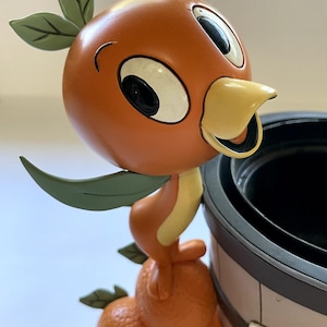 May include: A whimsical orange bird figurine with green leaves and a yellow beak. The bird is perched on a cluster of orange fruit. A decorative flower pot is in the background. The figurine is a collectible item.