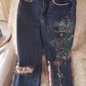 May include: Dark wash denim jeans with floral embroidery and patches. The jeans feature pink lace trim on the pockets and decorative patches with floral patterns. The embroidery includes green vines and red flowers.