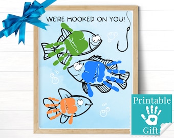 Könnte beinhalten: Ein gerahmter Kunstdruck auf hellblauem Hintergrund mit dem Text WE'RE HOOKED ON YOU! und drei Fischillustrationen. Die Fischkörper bestehen aus grünen, blauen und orangefarbenen Handabdrücken mit schwarzen Umrissen und einem Angelhaken.