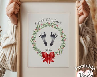 May include: A framed print featuring a watercolor wreath of green leaves and red berries surrounding two black baby footprints. The text reads My 1st Christmas and 2026, with a large red bow at the bottom of the wreath. The frame is light wood.