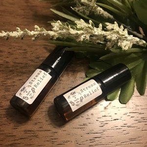 Könnte beinhalten: Zwei kleine Roll-on-Fläschchen aus bernsteinfarbenem Glas mit schwarzen Verschlüssen, jeweils mit einem weißen rechteckigen Etikett, das den Text Stress Relief und den Markennamen oil & herb co. in einem botanischen Kranz zeigt.