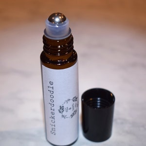 May include: A brown glass roller bottle with a silver roller ball. The bottle has a white label with the text "Snickerdoodle" and a black and white illustration of a wreath with the text "Oil & Herb". The bottle is sitting on a white surface.
